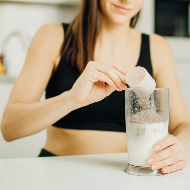 woman pouring protein powder into glass smoothie black tank top exercise clothes