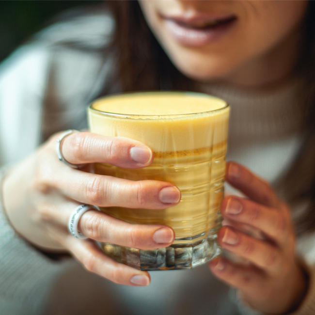 woman drinking yellow turmeric tea in glass blue sweater brown hair