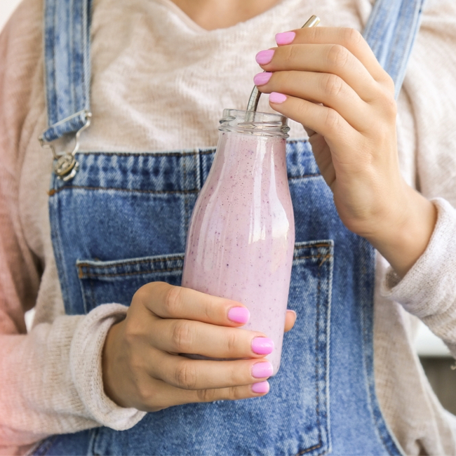 woman holding pink berry smoothie in glass with straw overalls tan shirt pink nails