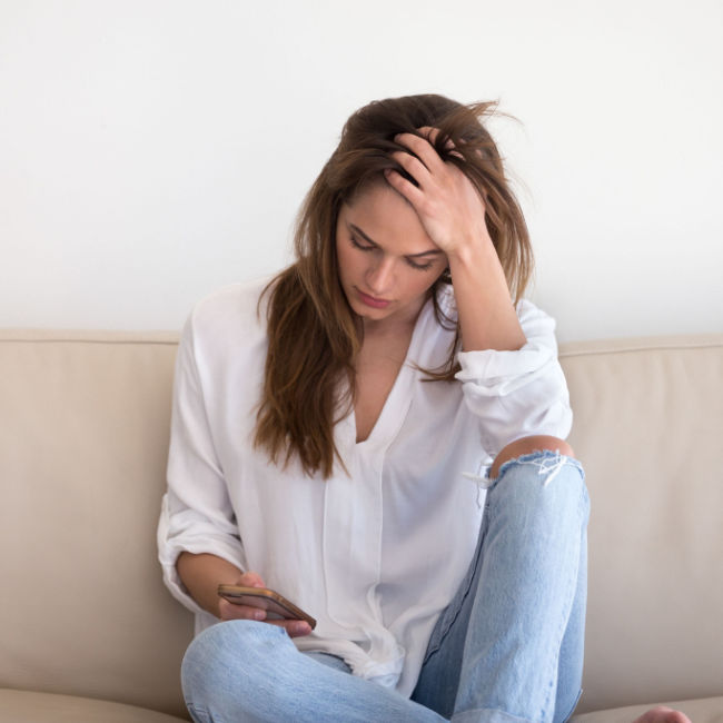 woman sitting on couch looking at phone holding forehead with upset expression