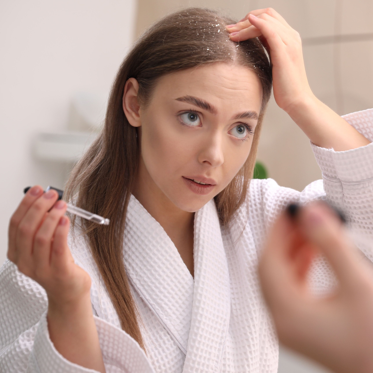 woman applying scalp oil parted brunette hair touching scalp serum applicator white robe bathroom mirror