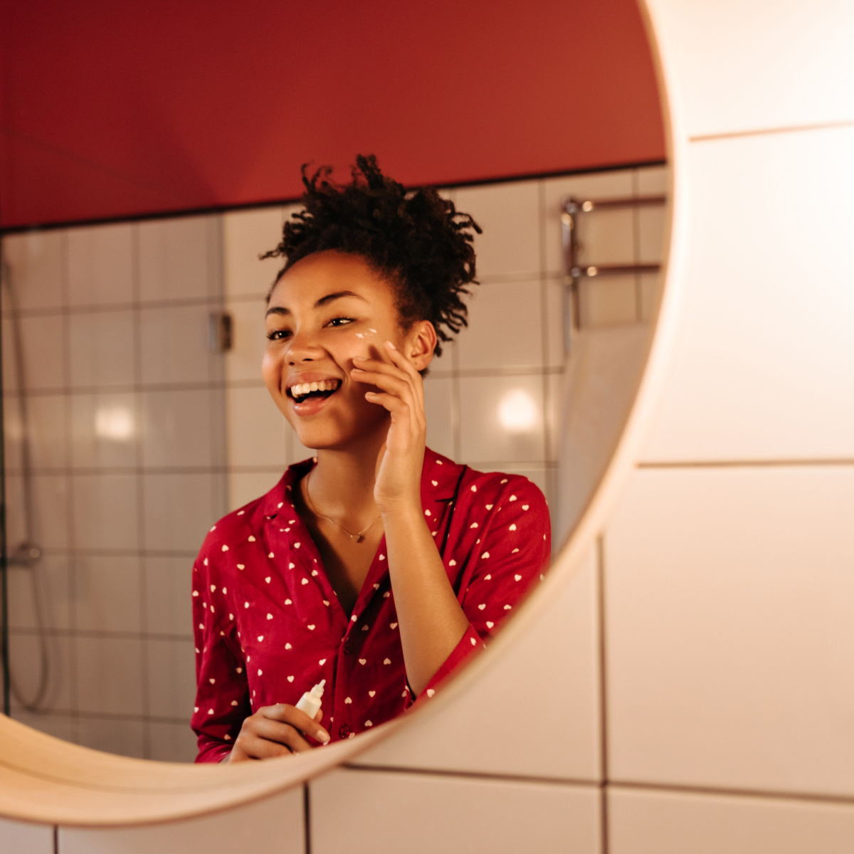 woman applying eye cream sunscreen to face glowing skin before bathroom mirror hair up red shirt