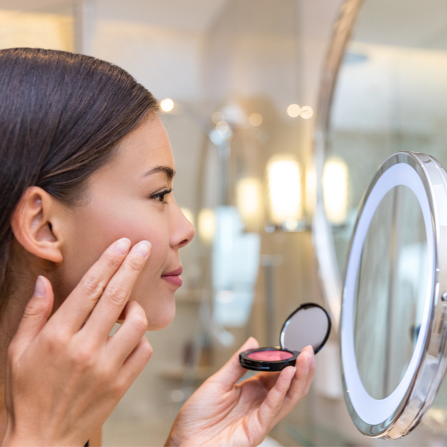 woman applying cream blush pink with fingers in front of bathroom mirror