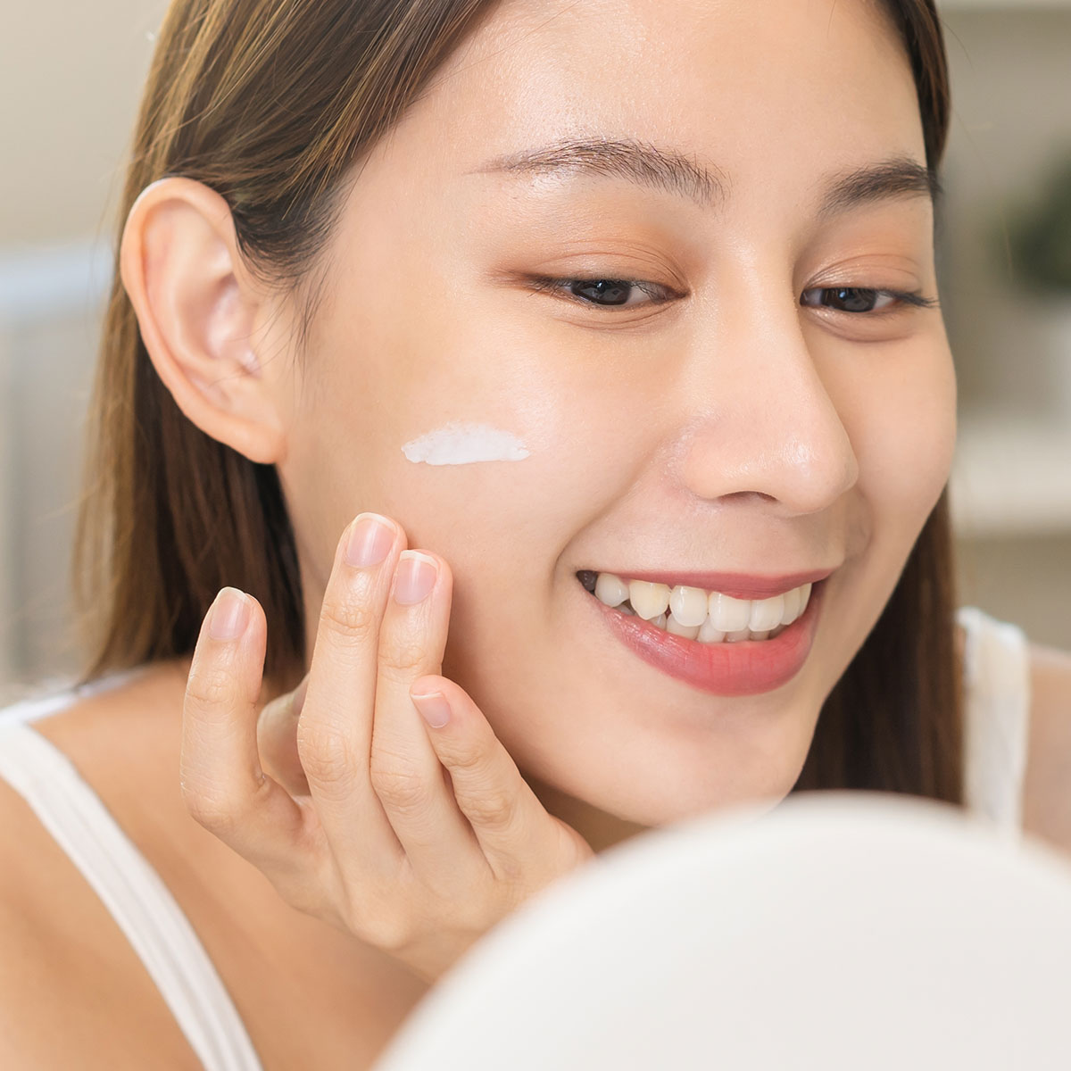 young woman applying sunscreen to cheek before white mirror smiling