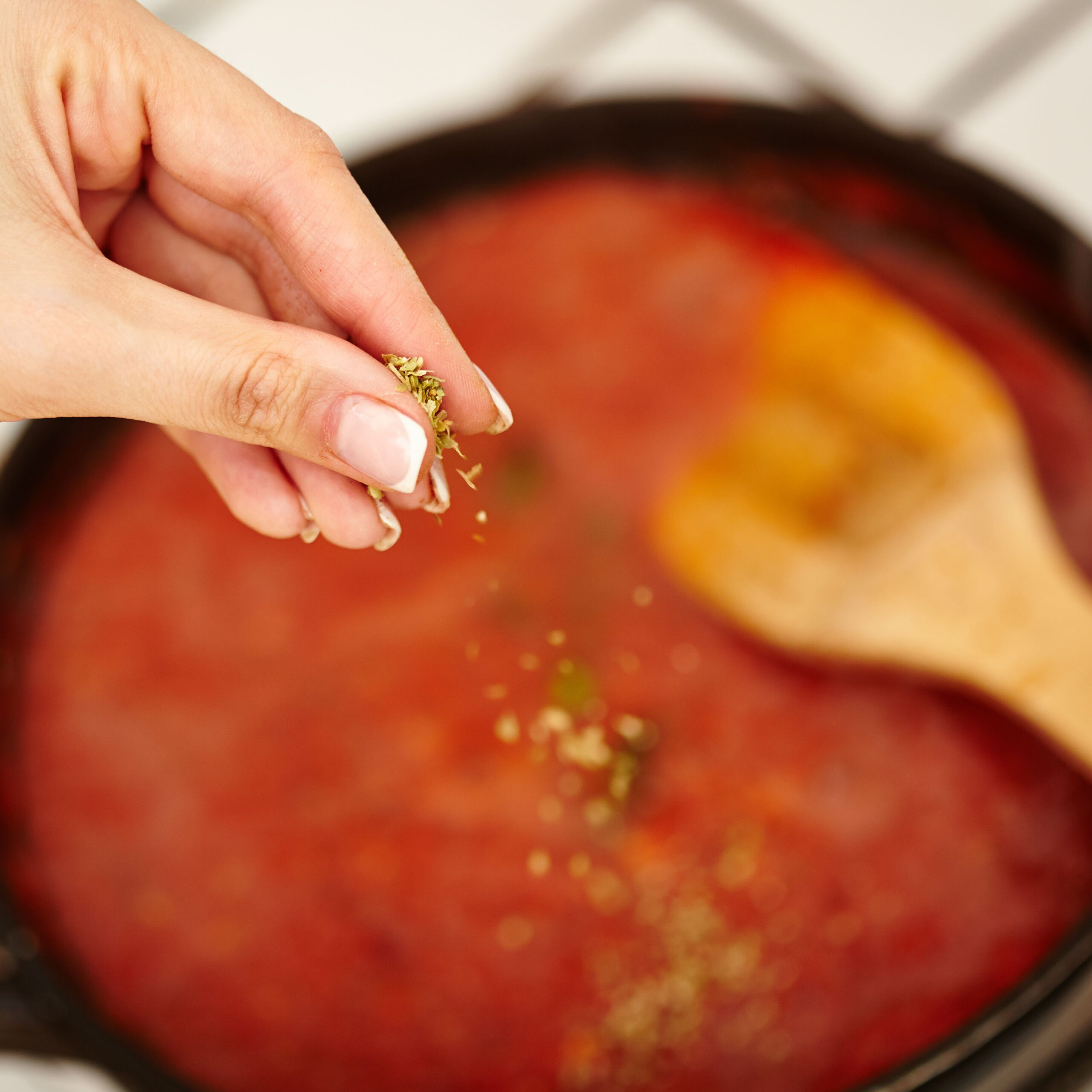 hand sprinkling herbs onto pan of pasta sauce